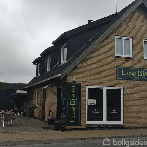 En gul murstensbygning med skiltet "Læsø Bistro" på facaden. Der står borde og stole opstillet udendørs på en flisebelagt terrasse. Bygningen er omkranset af en skyet himmel.