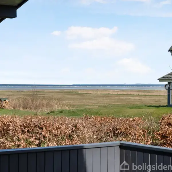 En havelampe står stille på en terrasse med udsigt over en flad, grøn eng mod havet. En række huse ses i baggrunden under en blå himmel.
