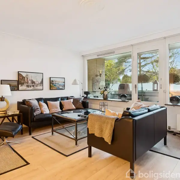 Sort læderstuesæt med sofabord ovenpå træparketgulv i lys, moderne stue. Store vinduer slipper naturligt lys ind. Dekorationer og lamper skaber en hyggelig atmosfære.