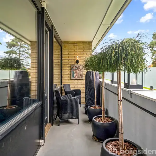 Balkon med sorte kurvemøbler og flere pottetræer, placeret op ad en murstenvæg med udsigt til træer og blå himmel. Ingen tekst er synlig.