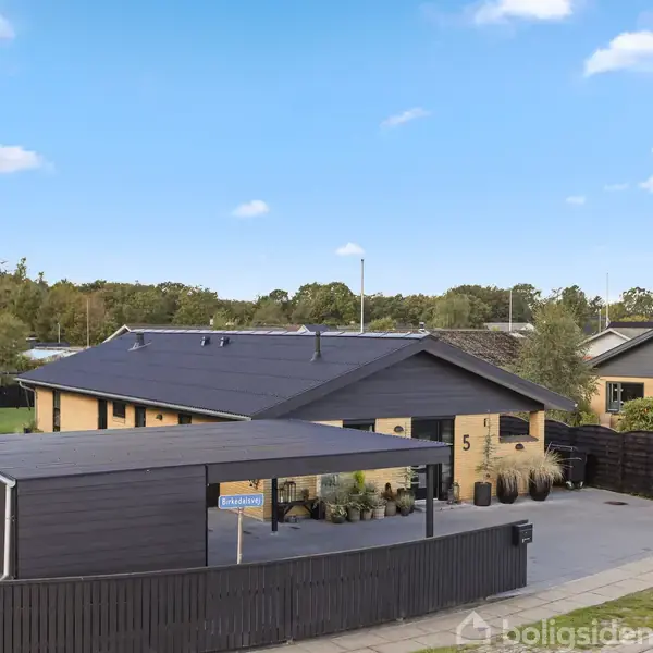 En gul murstensvilla med sort tag og nummeret "5" på facaden, står på en velholdt grund. En sort carport og indkørsel er synlige. Omgivet af grønt og blå himmel.