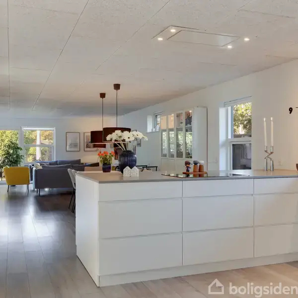 Et moderne køkken med hvide skabe og en køkkenø. Rummet er lyst med trægulv og åbner op mod en stue med sofagruppe og lys indretning. Vægur viser klokken 9.