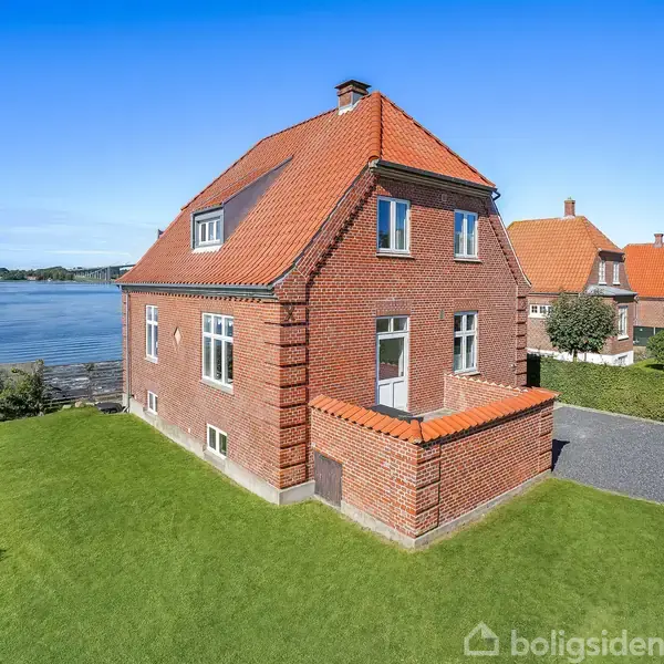 Rødt murstenshus med rødt tegltag står på en velholdt græsplæne ved siden af en grusvej. I baggrunden ses en sø og andre lignende huse langs vandet.