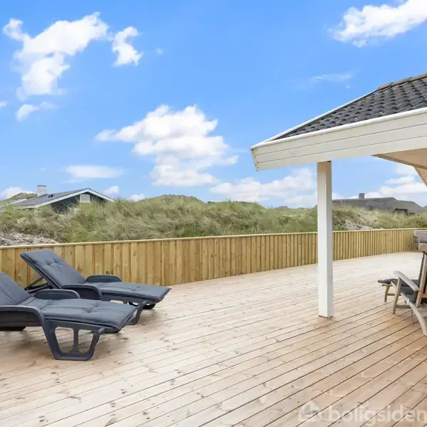 To liggestole står på en stor træterrasse ved et hus med overdækket område. Baggrunden viser en klit med græs og en blå himmel med skyer.