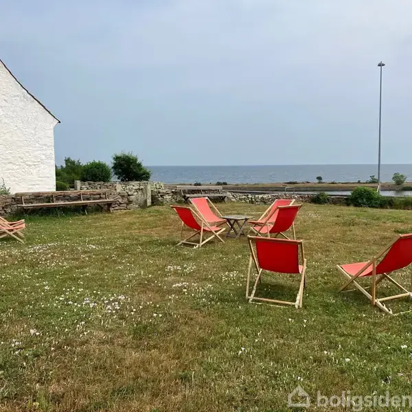 Røde klapstole står på en græsplæne ved siden af et hvidt hus nær vandet. En stenbænk og lavendelbuske ses ved siden af.