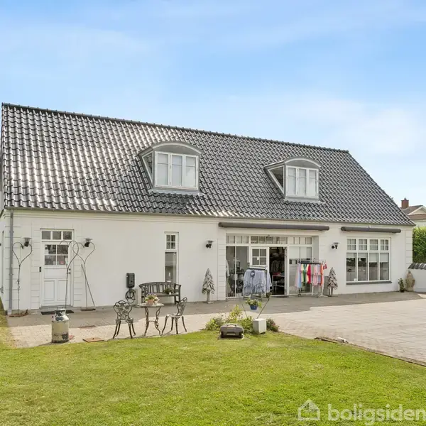 En hvid étplans villa med mørkt tegltag og to kviste; i haven står havemøbler på en flisebelagt terrasse omgivet af velplejet græsplæne under en klar blå himmel.