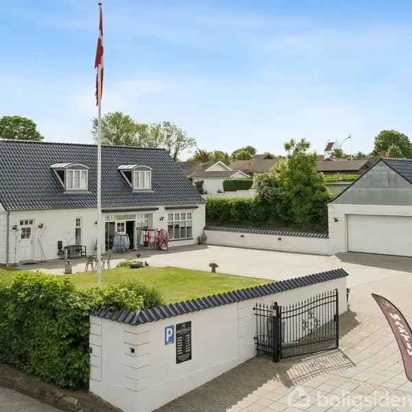 Hvidt hus med mørkt tag står foran flisebelagt indkørsel. Til venstre ses en flagstang med dansk flag. Til højre er en garage. Grønne hække omkranser ejendommen. Parkeringsskilt ved indgang.