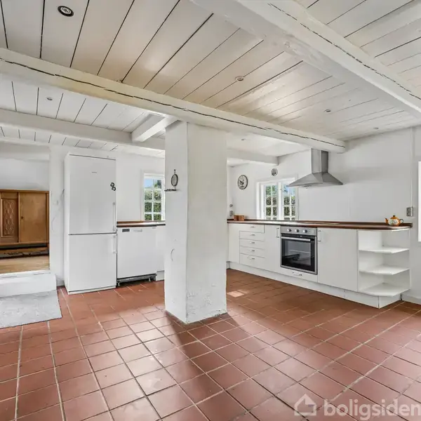 Køkken med hvidt interiør og terracotta-fliser, køleskab og ovn. Store vinduer giver udsigt til have. Loftet har synlige bjælker, og et skab ses i baggrunden.