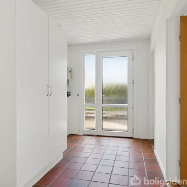 En lys entre med røde fliser, hvide skabe, og en glasdør der fører ud til en grøn udsigt udenfor.