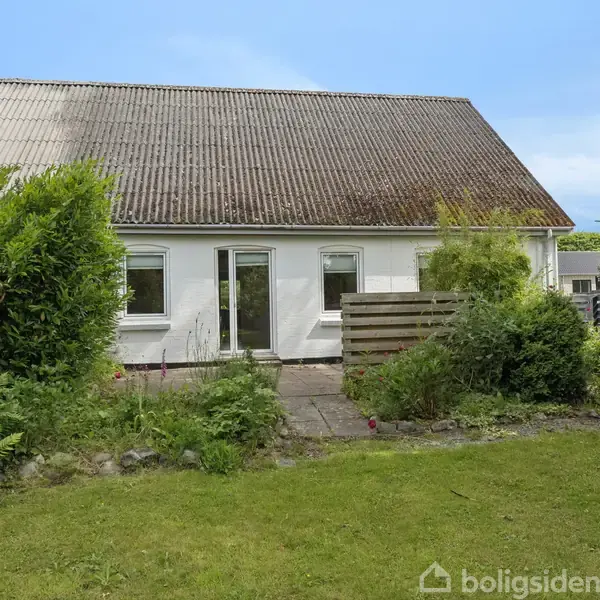 Et hvidt hus med skrånende tag ses fra en have med græs og buske. Der er en terrasse foran huset med en træafskærmning ved siden af.