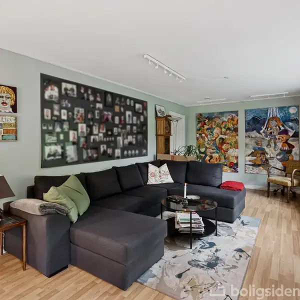 Stor, mørkegrå sofa står i lys stue med trægulv. Væggene har kunstværker og fotografier. Store vinduer slipper lys ind. To små borde med dekorationer fuldender indretningen.