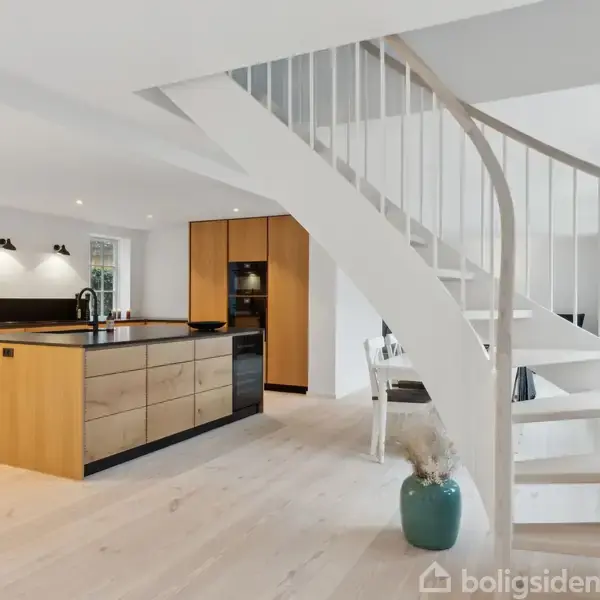 Spiraltrappe står i et rummeligt, lyst køkken med træskabslåger og midterø. Store vinduer giver naturligt lys, og en vase med dekorative grene står nær trappen.