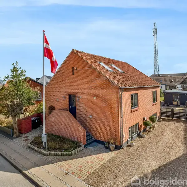 Rød murstenshus står på en hjørnegrund med dansk flag i flagstang foran. Omgivet af en have med grus indkørsel, ligger huset i et boligkvarter med blå himmel som baggrund.