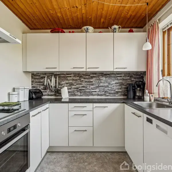 Et hvidt køkken med sorte bordplader har moderne apparater og en grå flisevæg. Solen skinner gennem et stort vindue, hvilket giver rummet naturligt lys.