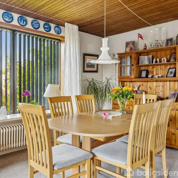 Et rundt spisebord med seks stole står i en spisestue med trævægpaneler. En stor reol i træ er fyldt med nipsgenstande. Vindue med persienner giver udsigt til haven.