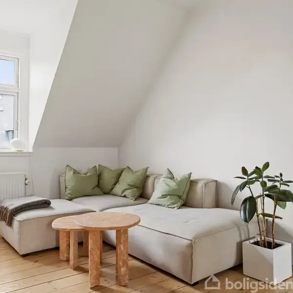 Sofa med grønne puder står i hjørnet af et lyst rum, ved siden af et lille rundt bord og en plante. En radiator og et vindue ses i baggrunden.