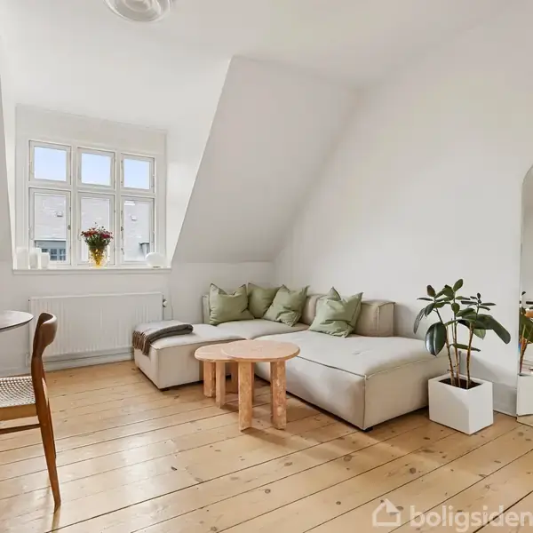 En stue med en lys sofa, pyntet med puder, står ved et stort vindue. Et rundt sofabord og en plante supplerer rummet. En spisebord står til venstre. Ingen tekst.