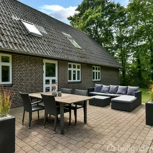 En terrasse med ude møbler, herunder et bord og stole, står foran en murstenshus. Der er store plantekasser ved siden af sofaen, omgivet af træer og græsplæne.