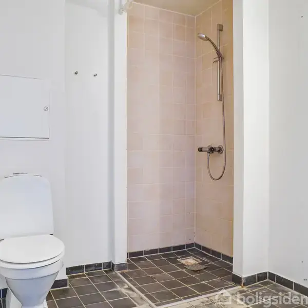 Toilet står stille i et badeværelse. Ved siden af er der en bruser med flisevæg. Rummet er tomt og enkelt indrettet.