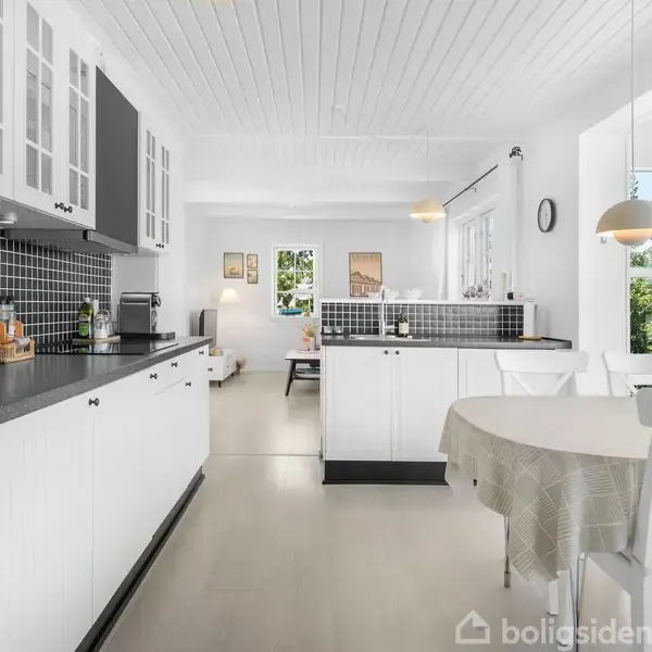 Køkken med sorte og hvide skabe, indbygget ovn og sort flisebaggrund. Rundt spisebord foran vindue, der slipper naturligt lys ind. Åben forbindelse til spise- og opholdsstue.