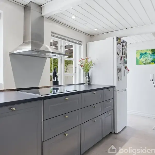 Et moderne køkken med grå skuffer, metalemhætte og en hvid køleskab ved siden af. Blomster står ovenpå skufferne, og et farverigt maleri hænger på væggen.