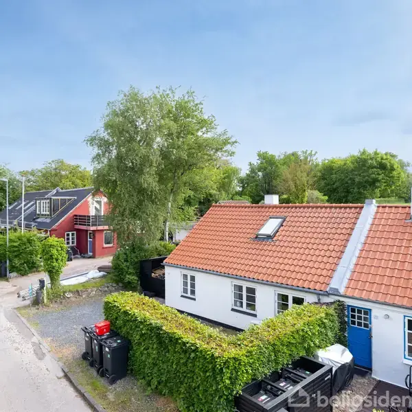Et hvidt hus med rødt tegltag står langs en stille villavej. En hæk omkranser haven, mens en rød bygning er synlig i baggrunden blandt frodige træer.