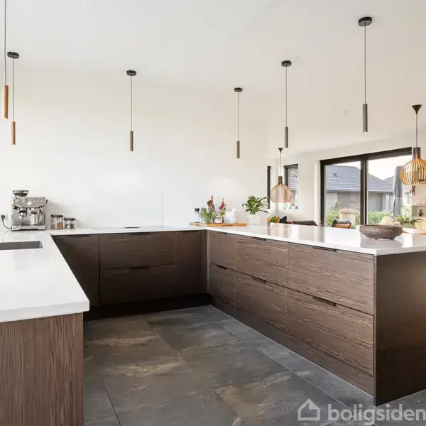 Køkken med moderne design; store, mørke skabe under hvide bordplader; hængelamper over bordpladen. Lys strømmer ind gennem store vinduer. I baggrunden ses et spiseområde med lamper.