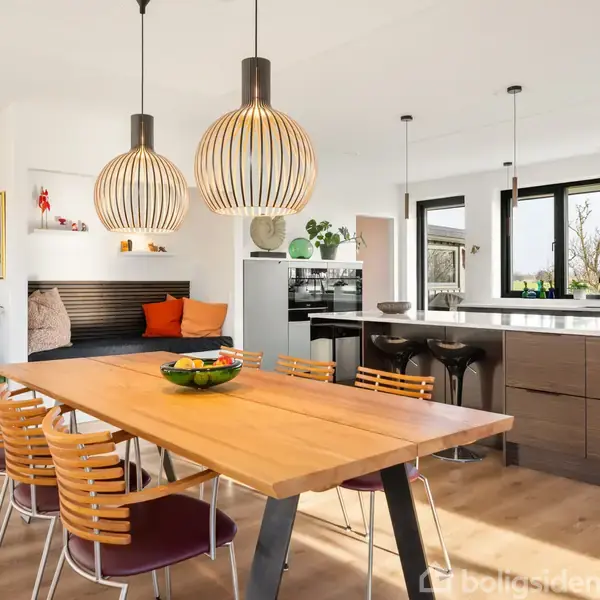 En træspisebord dekoreret med en skål frugt står under moderne lamper, omgivet af stole i et lyst køkken/alrum med store vinduer og en indbygget bænkesæde ved siden af planter.