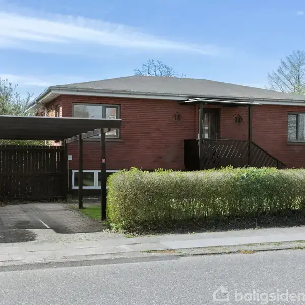 En etplans murstensvilla med et fladt tag står stille. En hækkant omkranser huset, og en carport er til venstre. Boligsiden vandmærke ses i nederste højre hjørne.