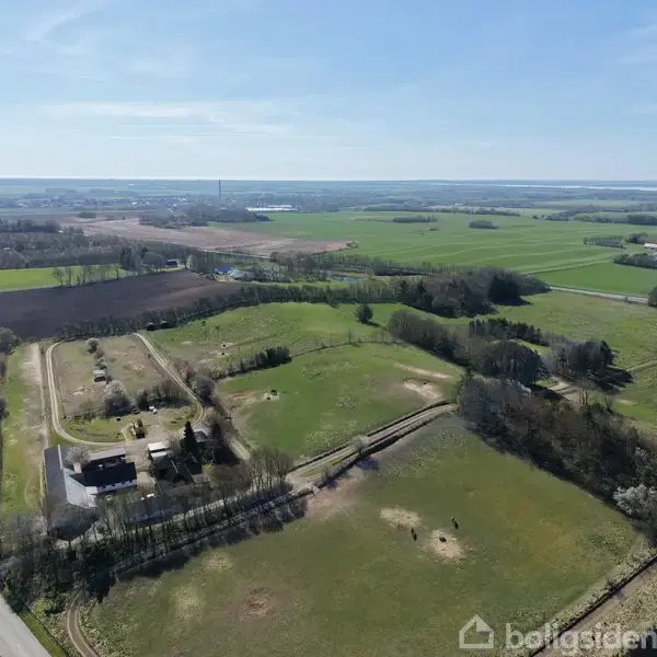 Grønne marker strækker sig over et fladt landskab, hvor et par bygninger ligger spredt. Området er omkranset af skove og veje, under en blå, skyfri himmel.