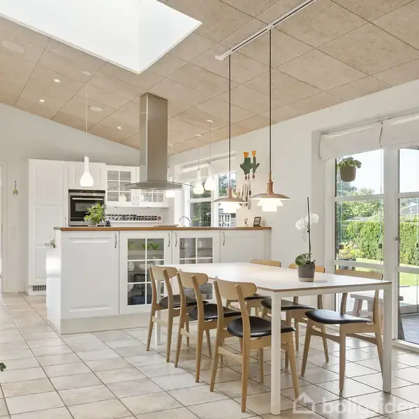 Et lyst og moderne køkken-alrum med ild på vægge og loft. Et træbord med stole står centralt. Store vinduer fører ud til en grøn have.