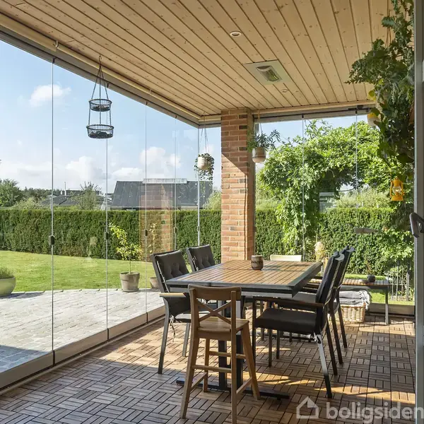 Et bord med stole står i en glasinddækket terrasse, omgivet af grønne buske og en velplejet græsplæne. Over dækker et loft af træ.