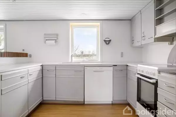 Hvidt køkken med moderne skabe og skuffer, ingen aktivitet, centralt vindue med udsigt til natur, trælaminatgulv, opvaskemaskine og ovn.