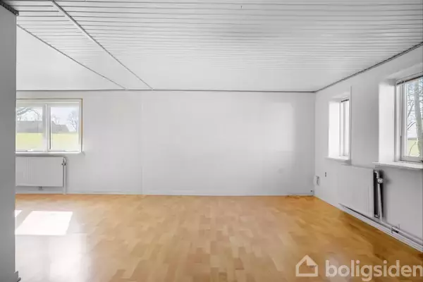 Rummet har lyse trægulve og hvide vægge. Tre vinduer lader dagslys strømme ind, med en radiator under hvert vindue. Værelset fremstår tomt og rummeligt.