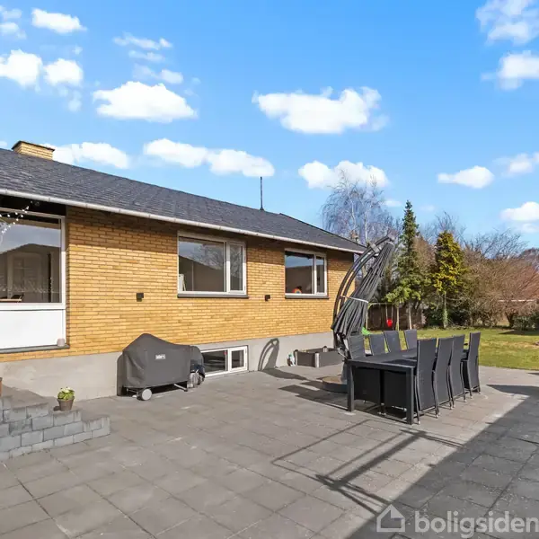 En gulstensvilla står i en have med en stor flisebelagt terrasse. Terrassen har havemøbler og en grill. Haven er omgivet af træer og buske, mens himlen er blå med få skyer.