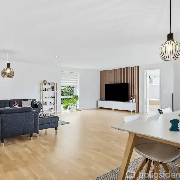 Stue med blå sofa centralt placeret, tv hængende på væggen, lys trægulv, spisebord i forgrunden og store vinduer der lukker dagslys ind. To moderne loftlamper.