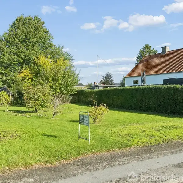 Et skilt står i en velplejet have, omgivet af grønne buske og græs, foran et hvidt hus med rødt tag. Skiltet har teksten: "Til Salg" og "RealMæglerne."
