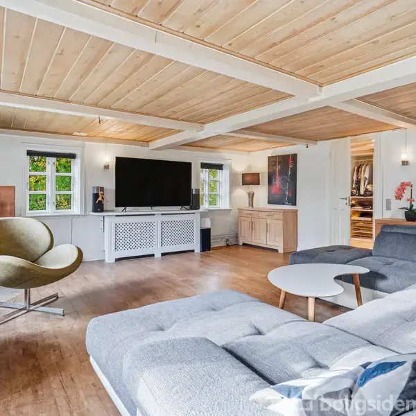 En moderne stue med en stor grå sofa, en rund grøn lænestol og et rundt sofabord. Stuen har trægulv, træloft, stor fladskærms-tv og små vinduer.