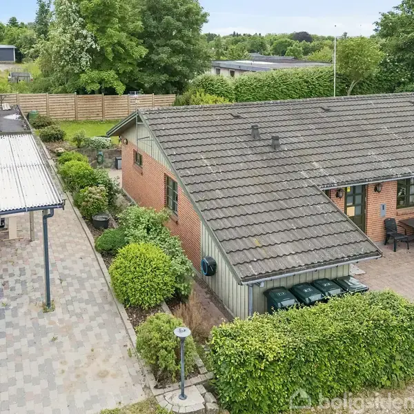 En murstensbygning med gråt tag i et villakvarter. Til højre ses en terrasse med havemøbler, og til venstre står en carport. Omkring er der grøn beplantning.
