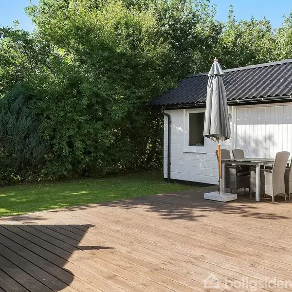 Træterrasse med borde og stole står foran et hvidt hus med sort tag. En parasol dækker spiseområdet. Omkranset af grønne buske og træer.