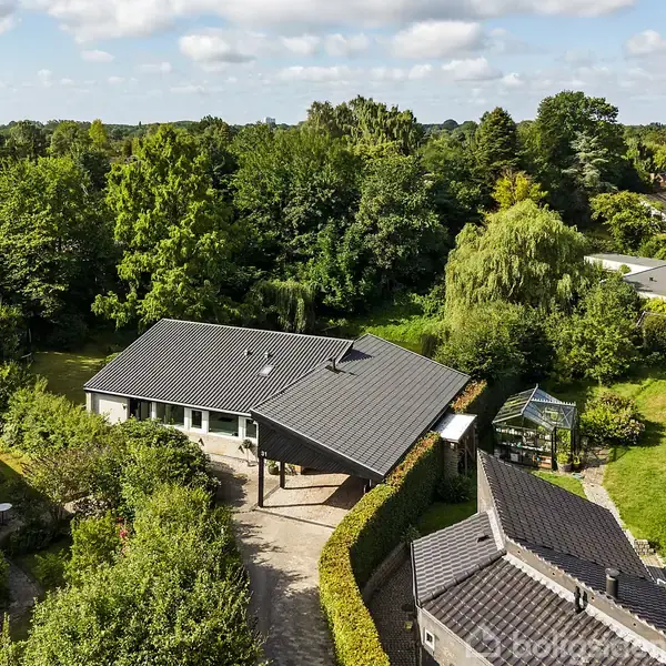 Et moderne hus med mørkt tag ligger omgivet af frodig grøn vegetation. Huset har en indkørsel med brosten og en velplejet have. Området er fyldt med tæt skov og åbne græsarealer.