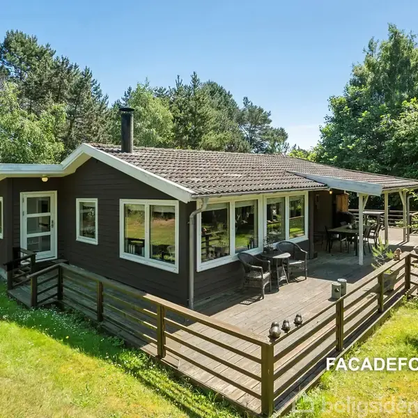 Et træhus med en stor terrasse har udendørs møbler stående. Huset er omgivet af en grøn græsplæne og tætte træer. Tekst: "FACADEFORSLAG".