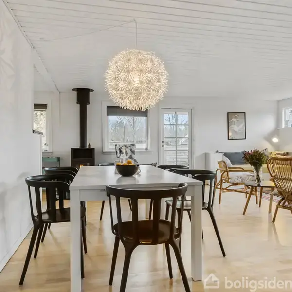 Spisebord med sorte stole står i et lyst rum med trægulv. En stor, kugleformet lampe hænger over bordet. I baggrunden ses en brændeovn, sofa og lænestol.
