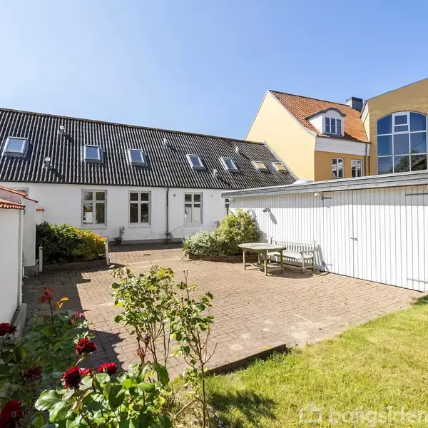 Hus med sorte tagsten og flere døre åbner op til en gårdhave med flisebelægning, bord og stole. Omkring er græs og blomsterbede. Klar himmel i baggrunden.