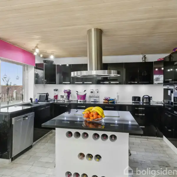 Et moderne køkken med sorte skabe og grå bordplader. Et stort spisebord med frugter i midten og indbygget vinreol. Lysindfald gennem vinduer med pink gardiner.