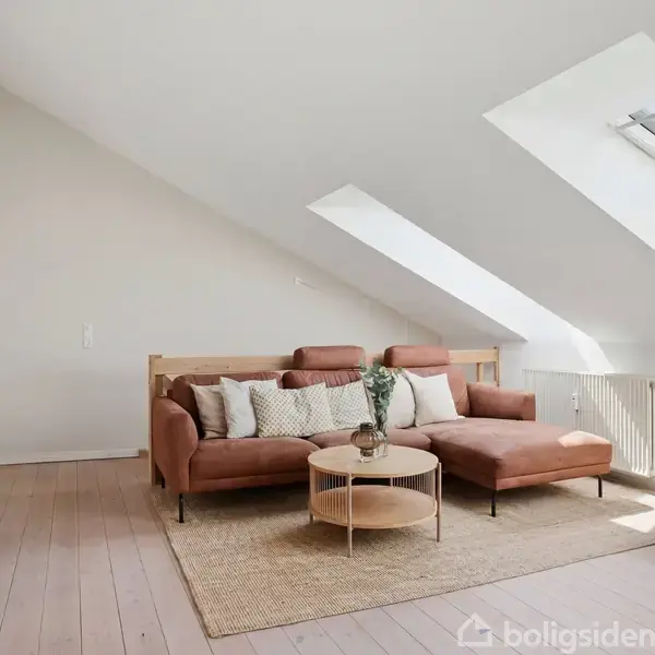 Brun sofa med puder står i skråt loftsrum, sollys strømmer ind gennem ovenlysvinduer. Et rundt sofabord står foran sofaen, ved siden af et lille, hvidt skab med blomster.