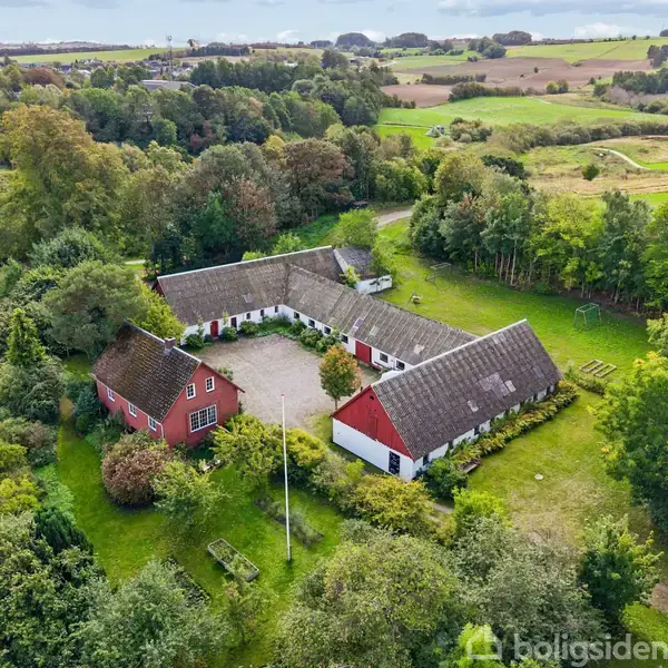 Landejendom set fra oven, med bygninger der danner en hesteskoform omkring en gårdsplads. Omgivet af marker og skove, fremhæves rødmalede og hvidmalede bygninger mod en frodig, grøn landskab.