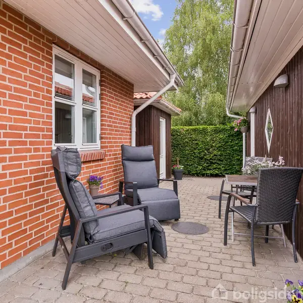 To havestole står på en terrasse med fliser mellem to bygninger i røde mursten og brune træpaneler. Der er en grøn hæk i baggrunden og et cafébord med sorte stole.