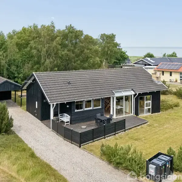 Et sort træhus ligger i en grøn have med en grusvej foran. Baggrunden viser flere huse og et kig til kysten.