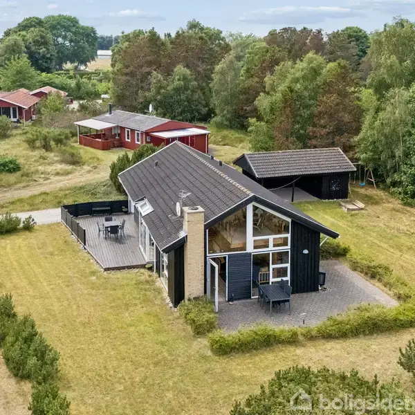 Et sommerhus med sort træfacade og terrasse står i et grønt område omgivet af træer. Der er en mindre rød bygning og en garage i baggrunden.